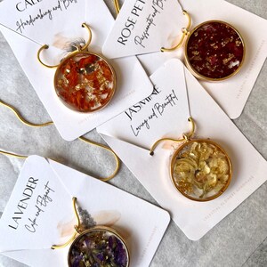 May include: Four gold necklaces with round pendants featuring pressed flowers and gold flakes. Each pendant has a different flower type and color, including orange, red, white, and purple. The necklaces are displayed on white cards with text describing the flower type and its meaning.