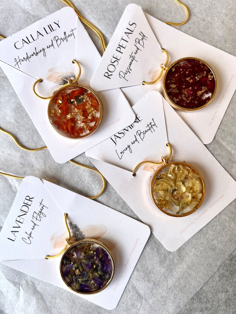 May include: Four gold necklaces with round pendants featuring pressed flowers and gold flakes. Each pendant has a different flower type and color, including orange, red, white, and purple. The necklaces are displayed on white cards with text describing the flower type and its meaning.