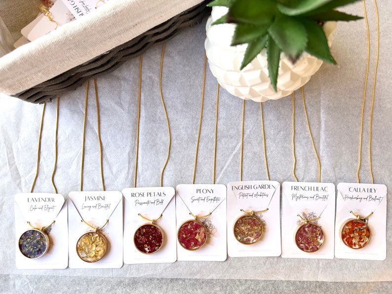 May include: Eight gold chain necklaces with round pendants featuring pressed flowers and gold flakes. Each pendant is labeled with a flower name and a short description, such as "Lavender, Calm and Elegant" or "Rose Petals, Passionate and Bold".
