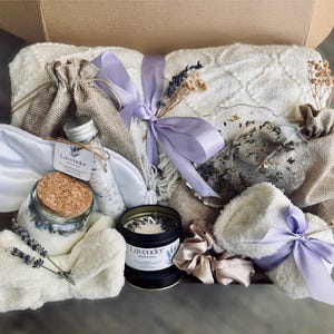 May include: A gift basket filled with lavender-themed self-care items, including a white blanket, a sleep mask, a jar of bath salts, a candle, and a pair of fluffy white towels.