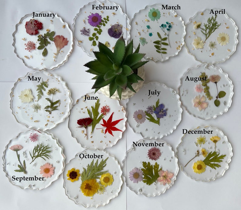 May include: Twelve round, clear resin coasters with a scalloped edge, each featuring pressed flowers and gold leaf. The coasters are labeled with the months of the year, January through December.
