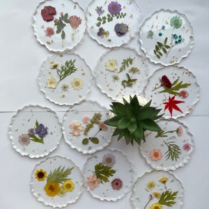 May include: Twelve round clear resin coasters with pressed flowers and leaves. The coasters have a scalloped edge and are arranged in a circle on a white surface.