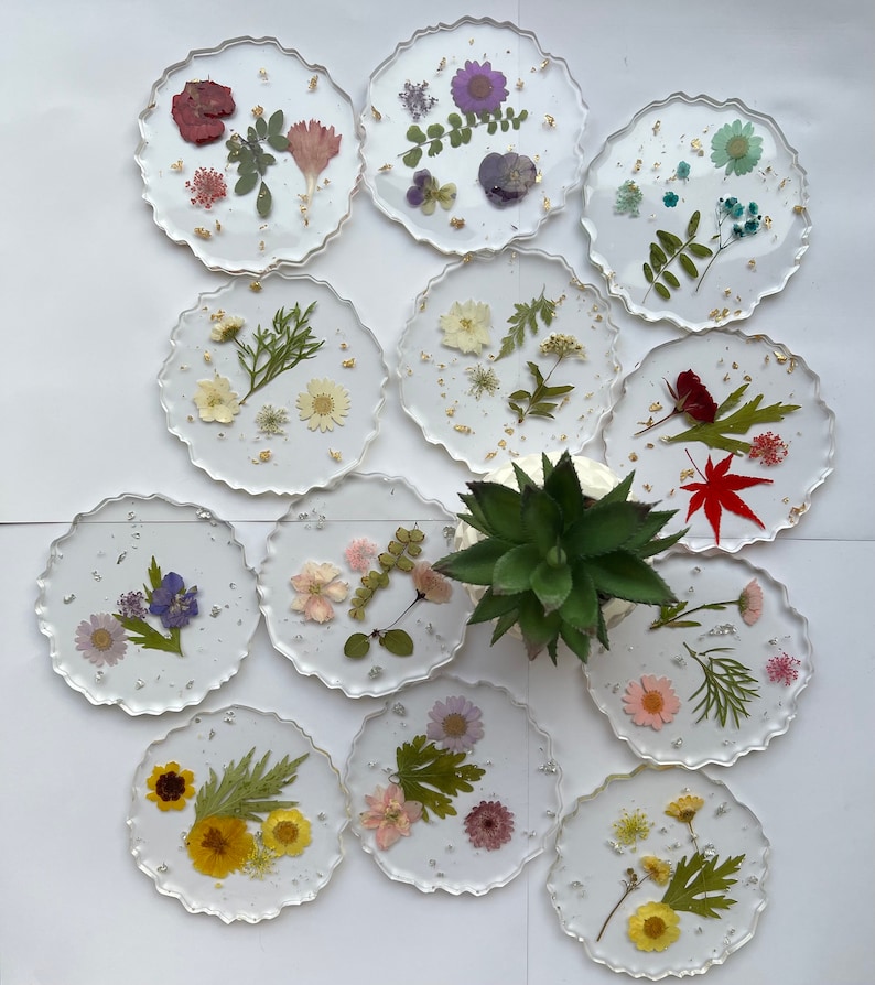 May include: Twelve round clear resin coasters with pressed flowers and leaves. The coasters have a scalloped edge and are arranged in a circle on a white surface.