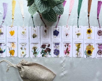 May include: Twelve bookmarks with pressed flowers and a small burlap bag. Each bookmark features a different month of the year, with the month name printed at the top. The bookmarks are decorated with pressed flowers and have a tassel attached to the top.