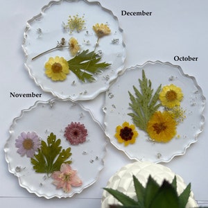 May include: Three round, clear resin coasters with pressed flowers and silver glitter. The coasters are labeled "November", "December", and "October".
