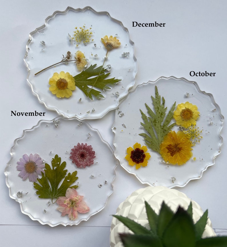 May include: Three round, clear resin coasters with pressed flowers and silver glitter. The coasters are labeled "November", "December", and "October".