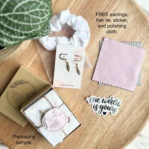 May include: A brown cardboard box with a white tissue paper interior, tied with twine. The box is labeled "Celestial Flowers Handmade". A white scrunchie, a pair of gold earrings, a pink polishing cloth, and a white sticker with the text "the world is yours" are also shown.