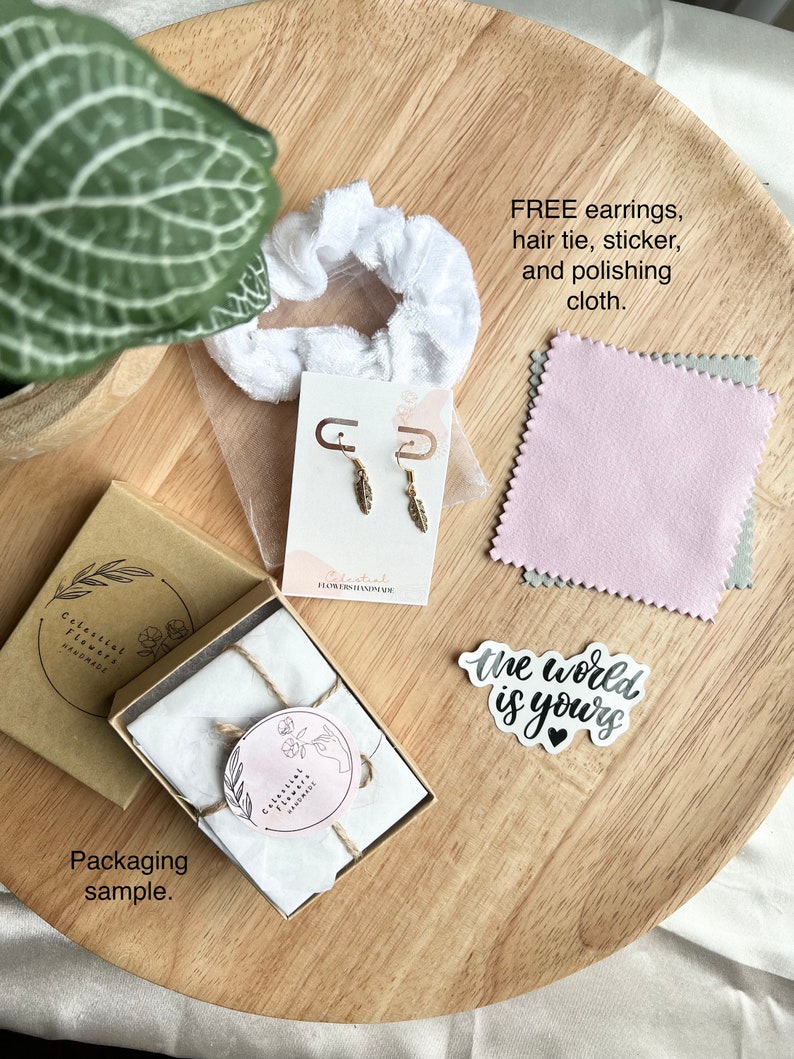 May include: A brown cardboard box with a white tissue paper interior, tied with twine. The box is labeled "Celestial Flowers Handmade". A white scrunchie, a pair of gold earrings, a pink polishing cloth, and a white sticker with the text "the world is yours" are also shown.