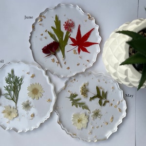 May include: Three clear resin coasters with pressed flowers and gold leaf. The coasters are labeled "April", "May", and "June".