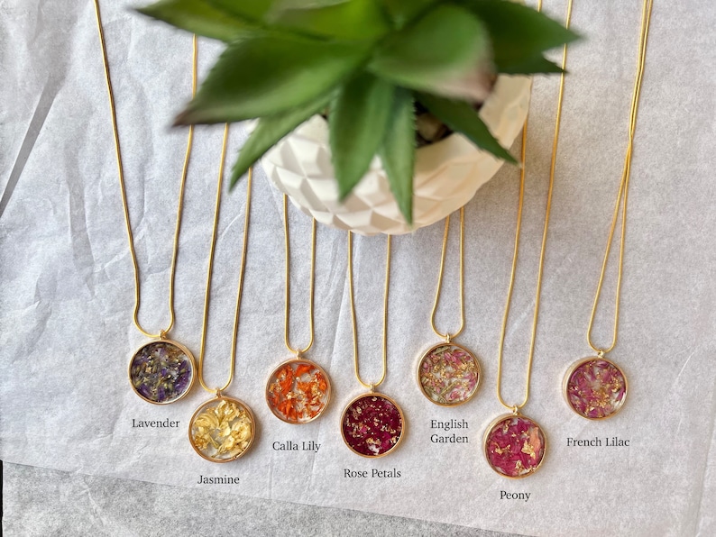 May include: Seven gold chain necklaces with round pendants containing pressed flowers and gold flakes. The pendants are labeled with the names of the flowers: Lavender, Jasmine, Calla Lily, Rose Petals, English Garden, Peony, and French Lilac.
