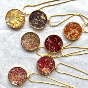 May include: Seven gold necklaces with round pendants featuring pressed flowers and gold flakes. Each pendant has a different color and type of flower.