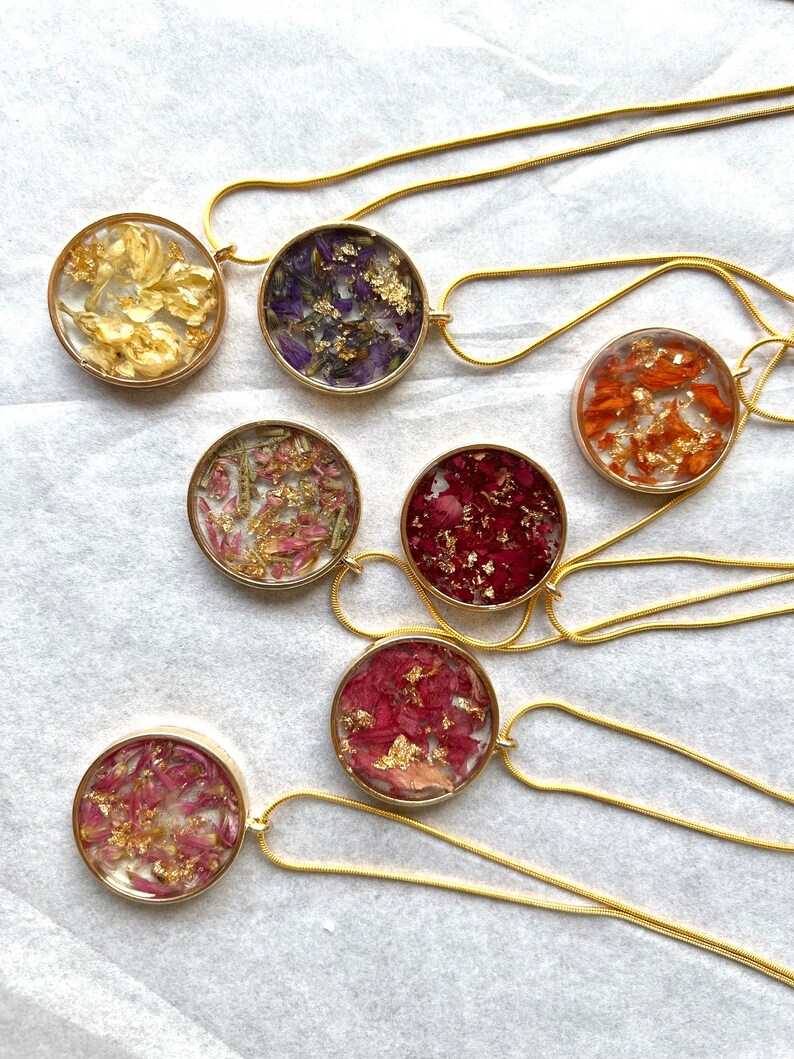 May include: Seven gold necklaces with round pendants featuring pressed flowers and gold flakes. Each pendant has a different color and type of flower.