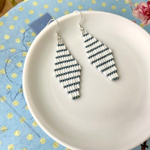 Könnte beinhalten: Ein Paar weiße und graue Perlen-Ohrringe mit einem Diamanten-Design. Die Ohrringe liegen auf einem weißen Teller mit blauen Punkten.