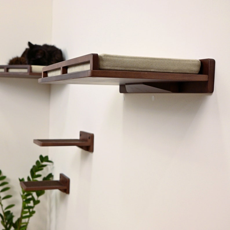 May include: Three wooden cat shelves mounted on a white wall. The top shelf has a beige cushion and is angled slightly forward. The middle shelf is a simple rectangular shape. The bottom shelf is also rectangular and is slightly angled forward.