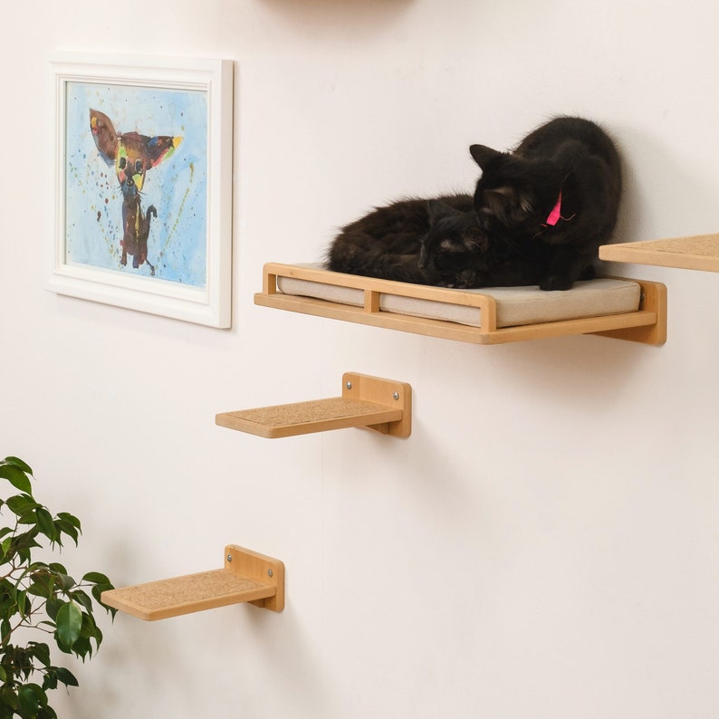 May include: A set of three wall-mounted cat shelves made of light wood. The top shelf has a soft cushion and two black cats are resting on it. The middle shelf is made of a textured material and the bottom shelf is made of smooth wood.