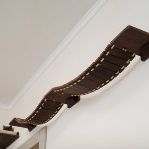 Op de afbeelding: Een gebogen houten kattenbrug met touwaccenten gemonteerd op een witte muur. De brug is gemaakt van donkerbruin hout en heeft een natuurlijk touw dat door de latten is geweven.