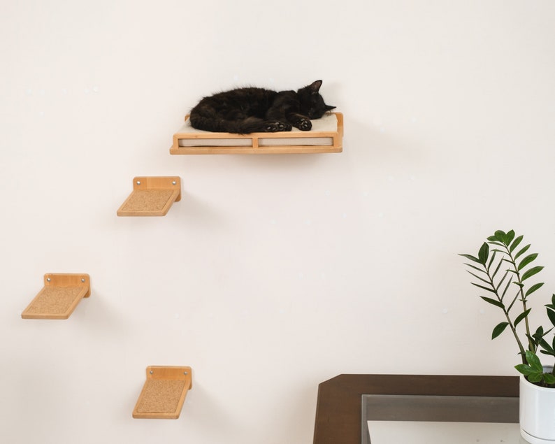 May include: A black cat rests on a wall-mounted cat shelf with a light-colored cushion. Below, three additional wooden cat steps with textured surfaces are mounted on the wall. The cat furniture is made of light-colored wood.