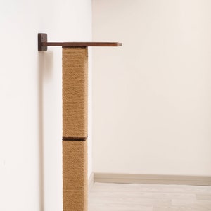 May include: A wall-mounted cat tree with a sisal scratching post and two wooden platforms. The platforms and mounting brackets are dark brown, contrasting with the light beige sisal and white wall. The cat furniture is designed for climbing and scratching.