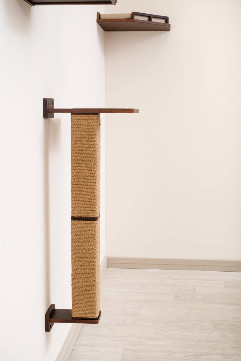 May include: A wall-mounted cat tree with a sisal scratching post and two wooden platforms. The platforms and mounting brackets are dark brown, contrasting with the light beige sisal and white wall. The cat furniture is designed for climbing and scratching.