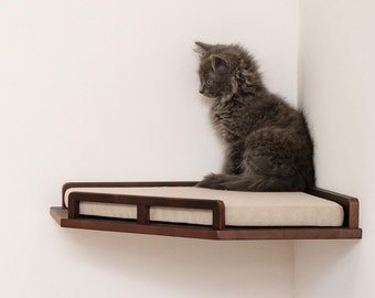 May include: A gray cat sits on a corner shelf bed for cats. The shelf is made of dark wood and has a beige cushion.
