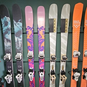 May include: A wall display of eight pairs of skis, with various colourful designs and brand names. The skis are mounted on a green wall with white trim. The brands include Armada, Mindbender, and Black Crows.