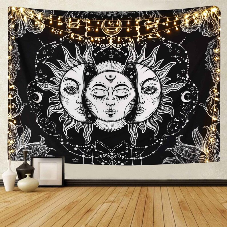 May include: Black and white tapestry featuring a sun and moon design with a celestial theme. The tapestry has a floral pattern around the edges and a string of lights hanging above it.