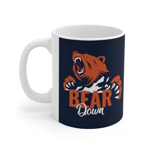 May include: White ceramic mug with a navy blue design featuring a roaring brown bear with claws, peeking out from behind a white surface. The text "BEAR Down" is printed below the bear in white and orange.