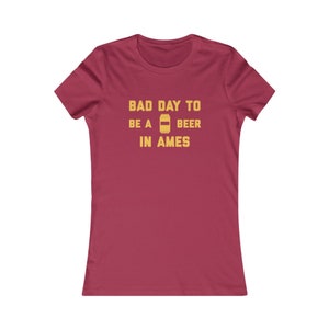 Può includere: Una maglietta da donna color granata con il testo "Bad Day To Be A Beer In Ames" in lettere gialle. Un'icona di lattina di birra gialla è inclusa nel testo.