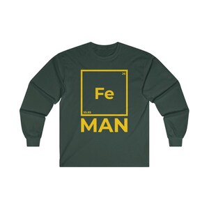 Puede incluir: Una camiseta de manga larga verde oscuro con un gr&aacute;fico cuadrado amarillo que presenta el s&iacute;mbolo qu&iacute;mico "Fe" para el hierro, el n&uacute;mero at&oacute;mico "26" y el peso at&oacute;mico "55,85". El texto "MAN" est&aacute; impreso debajo del cuadrado.
