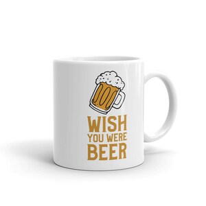 Può includere: Tazza in ceramica bianca con un'illustrazione di un boccale di birra in stile cartone animato e il testo "WISH YOU WERE BEER" in arancione.