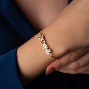 May include: A gold bracelet featuring five heart-shaped gemstones in shades of red, clear, and purple. The bracelet is worn on a wrist against a dark blue background. The gemstones are set in gold bezels.