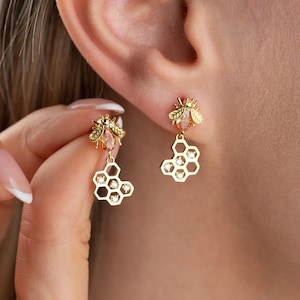 May include: Gold-toned bee earrings with a honeycomb design. Each earring features a small bee with a peach-colored gem and a dangling honeycomb shape with embedded clear crystals. The earrings are shown on an ear.