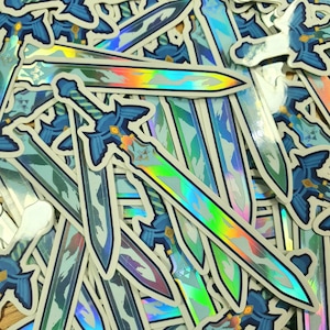 May include: A collection of holographic sword stickers with blue and silver details. The stickers feature a stylized sword design with a detailed hilt and blade, reflecting a rainbow of colors. The stickers are scattered across a surface.