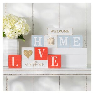 May include: Wooden block letters spelling "WELCOME HOME LOVE is in the air" in blue, white, and red with a gold heart and house graphic.