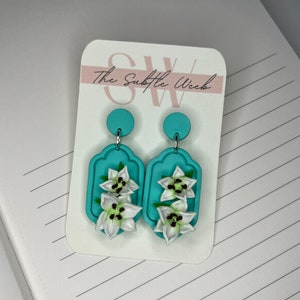 May include: A pair of turquoise blue earrings with white flower accents. The earrings are shaped like a tag with a rounded top and feature three white flowers with brown centers. The earrings are displayed on a white card with the text "The Subtle Week".