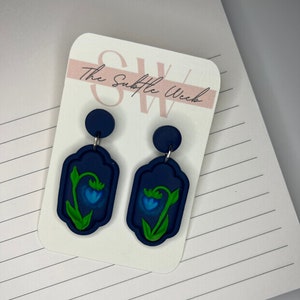 May include: A pair of blue and green floral earrings with a dark blue background. The earrings are on a white card with the text "The Subtle Week".