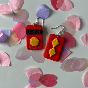 May include: A pair of red clay earrings with geometric designs. One earring features a yellow circle and the other features a yellow diamond shape.