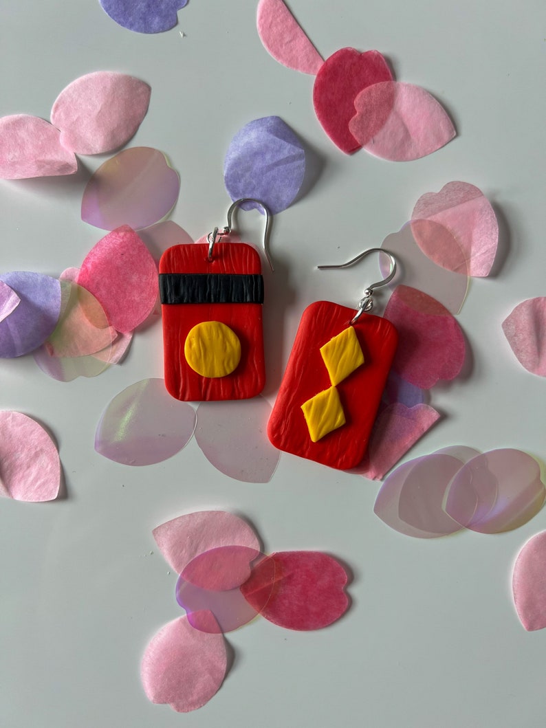 May include: A pair of red clay earrings with geometric designs. One earring features a yellow circle and the other features a yellow diamond shape.