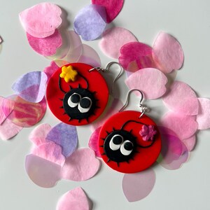 May include: A pair of red and black earrings featuring a cartoon character with large eyes and a star on top. The earrings are surrounded by pink and purple paper flower petals.