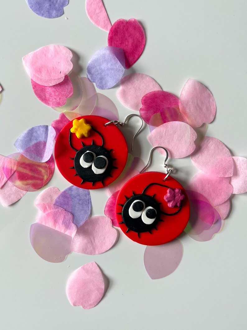 May include: A pair of red and black earrings featuring a cartoon character with large eyes and a star on top. The earrings are surrounded by pink and purple paper flower petals.
