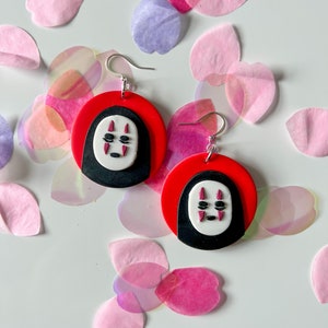 May include: A pair of red and black earrings with a white No Face character from the movie Spirited Away. The earrings are made of polymer clay and have silver hooks.