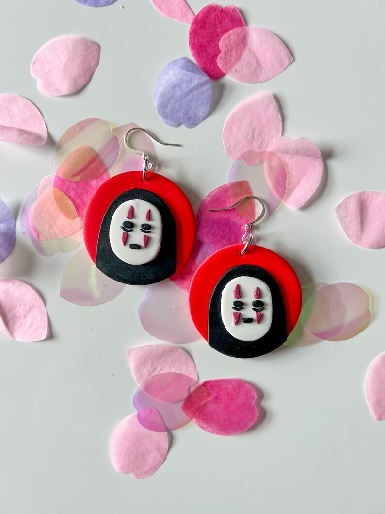 May include: A pair of red and black earrings with a white No Face character from the movie Spirited Away. The earrings are made of polymer clay and have silver hooks.