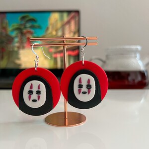 May include: A pair of red and black earrings with a white No Face character from the movie Spirited Away. The earrings are hanging from a gold stand.