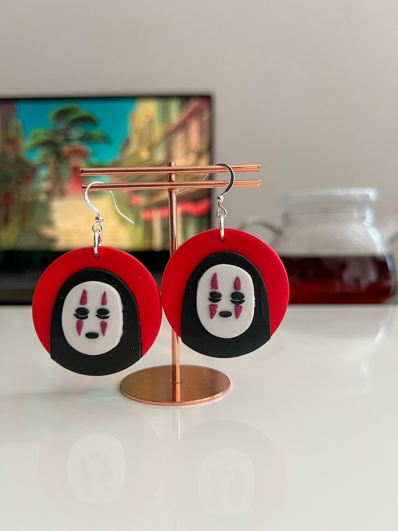 May include: A pair of red and black earrings with a white No Face character from the movie Spirited Away. The earrings are hanging from a gold stand.