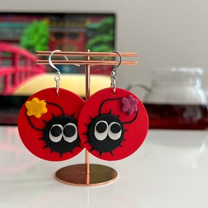 May include: A pair of red circular earrings with black and white eyes, a black outline, and a small yellow or purple flower on each earring.