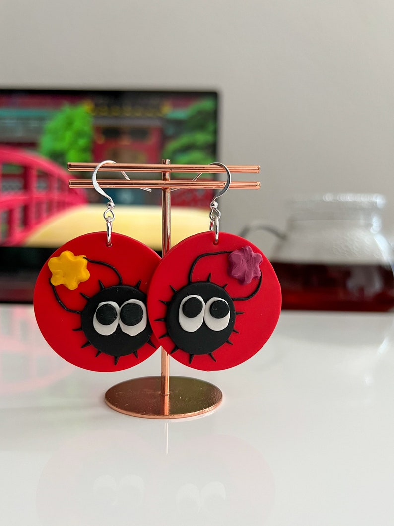 May include: A pair of red circular earrings with black and white eyes, a black outline, and a small yellow or purple flower on each earring.
