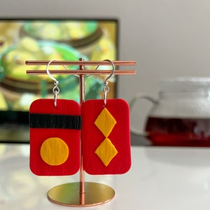 May include: A pair of red and yellow polymer clay earrings. One earring features a red rectangle with a black stripe and a yellow circle. The other earring features a red rectangle with two yellow diamonds.