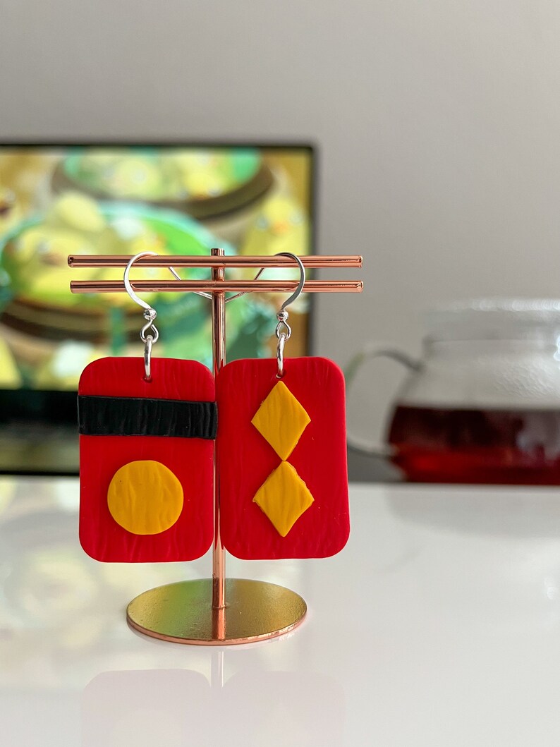 May include: A pair of red and yellow polymer clay earrings. One earring features a red rectangle with a black stripe and a yellow circle. The other earring features a red rectangle with two yellow diamonds.
