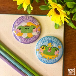 Op de afbeelding: Twee ronde, kleurrijke buttonspins met kikkerillustraties. Eén button is paars met de tekst "CERTIFIED BUFFOON", de andere lichtblauw met "CERTIFIED CLOWN". Beide hebben een cartoon kikkerontwerp.