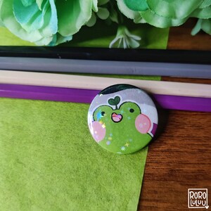 May include: A green frog with a pink nose and a green heart on its head. The frog is surrounded by a holographic glitter design. The background is a green and purple color scheme.